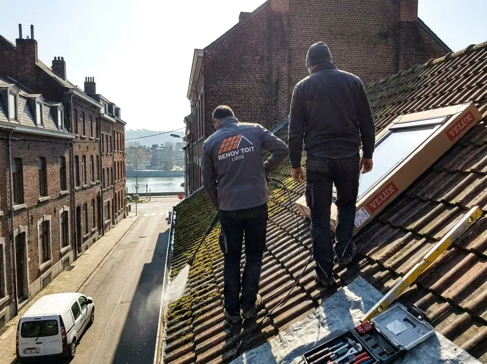 Deux couvreurs Renov'Toit Liège sur une toiture en pannes, fenêtre de toit prête à poser, rue descendant vers la Meuse